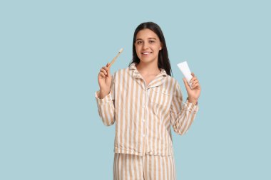 Mavi arka planda diş fırçası ve yapıştırıcısı olan pijamalı genç bir kadın.