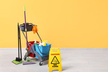 Temizlik malzemeleri ve sarı duvarın yanında uyarı levhası olan kapıcı vagonu.