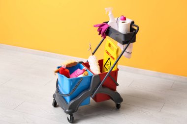 Temizlik malzemeleri ve sarı duvarın yanında uyarı levhası olan kapıcı vagonu.