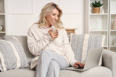 Evdeki kanepede dizüstü bilgisayar kullanan, kahve içen olgun bir kadın.