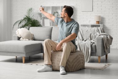 Klimalı uzaktan kumandalı genç adam beyaz tuğla duvarın yanındaki puf üzerinde oturuyor.