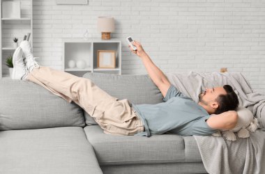 Klimalı uzaktan kumandalı genç adam beyaz tuğla duvarın yanındaki kanepede yatıyor.