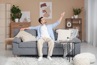 Klimalı genç adam bej duvarın yanındaki kanepede oturuyor.