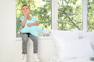 Yatak odasındaki pencere eşiğinde yastıklı düşünceli küçük çocuk.