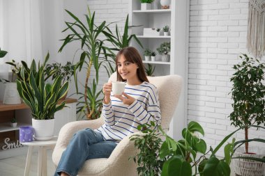 Evdeki çiçeklerin yanındaki koltukta oturan bir fincan çayla genç bir kadın.