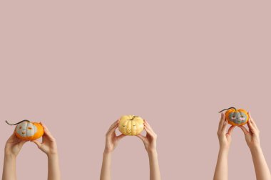 Yüzleri çizilmiş balkabaklarını tutan kadın elleri, pembe arka planda kil maskeler ve göz altı yamaları