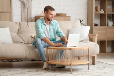 Genç adam odasındaki kanepede akıllı ev uygulamalı dizüstü bilgisayar kullanıyor.