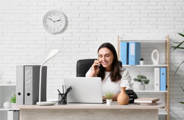 Modern dizüstü bilgisayarı ve çalışma alanında hava nemlendiricisi olan genç ve güzel bir kadın ofisinde telefonla konuşuyor.