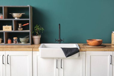 Beyaz seramik lavabo ve raf ünitesi mutfaktaki koyu mavi duvarın yanındaki ahşap tezgahın üzerinde.