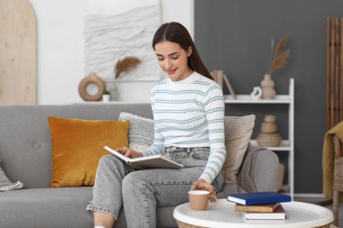 Evdeki kanepede çay içip kitap okuyan güzel, genç ve mutlu bir kadın.