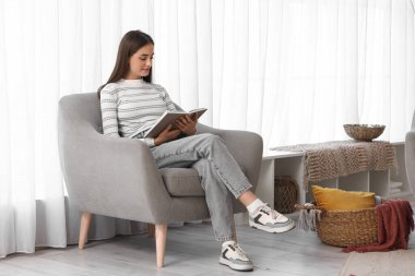 Evdeki pencerenin yanındaki koltukta kitap okuyan güzel, genç ve mutlu bir kadın.