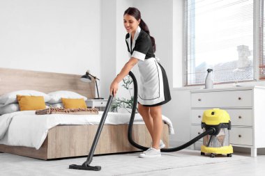 Genç oda hizmetçisi otel odasında halı süpürüyor.