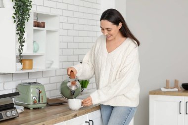 Güzel, genç, mutlu bir kadın mutfaktaki bardağa modern elektrikli çaydanlıktan sıcak su dolduruyor.
