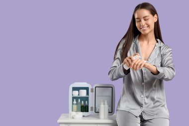 Masanın yanındaki mutlu genç kadın mor arka planda küçük bir kozmetik buzdolabı açtı.