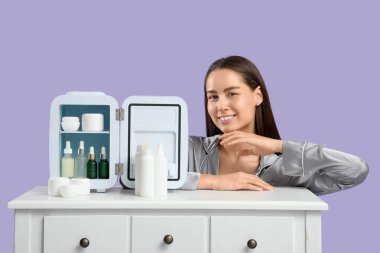 Masanın yanındaki mutlu genç kadın mor arka planda küçük bir kozmetik buzdolabı açtı.