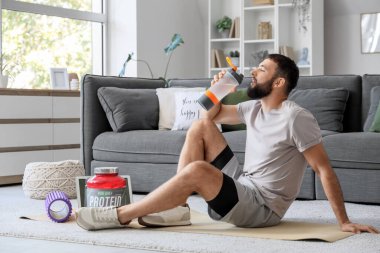 Sportif genç adam evdeki yoga minderinde protein içeceği içiyor.