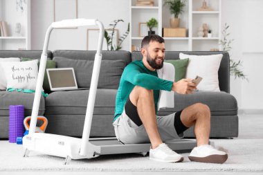 Cep telefonu ve havlusuyla evdeki koşu bandında oturan sportif genç adam.