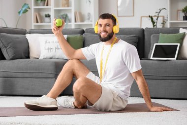 Kulaklıklı, elmalı ve mezuralı genç adam evde yoga minderinde oturuyor.