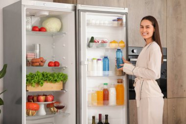 Genç bir kadın mutfaktaki temiz yiyeceklerle dolu açık buzdolabından su şişesini alıyor.