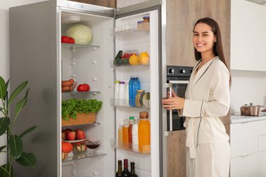Genç bayan açık buzdolabının yanında mutfaktaki taze yiyeceklerle dolu.