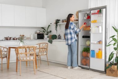 Genç bir kadın mutfaktaki yiyecek dolu açık buzdolabından taze elma alıyor.