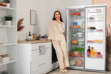 Genç bir kadın mutfaktaki açık buzdolabının yanında taze elma yiyor.