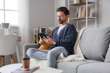 E-okuyuculu genç adam evdeki kanepede oturuyor.