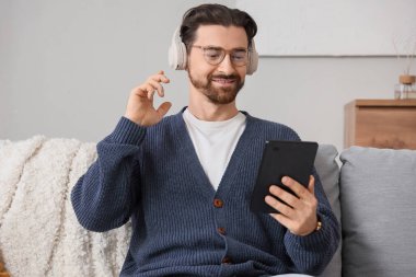 Kulaklıklı, e-okuyuculu genç adam evdeki kanepede oturuyor.