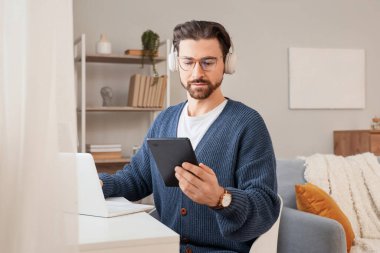 Kulaklıklı, e-okuyuculu genç adam evdeki masada dizüstü bilgisayar kullanıyor.