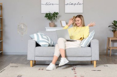 Dizüstü bilgisayarlı genç bir kadın evdeki gri koltukta sohbet ediyor.