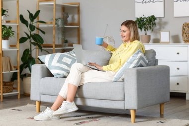 Evdeki gri koltukta dizüstü bilgisayar kullanan bir fincan çayla genç bir kadın.