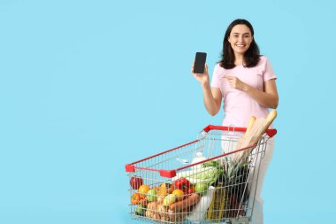 Mavi arka planda cep telefonunu gösteren alışveriş arabalı mutlu genç bir kadın.