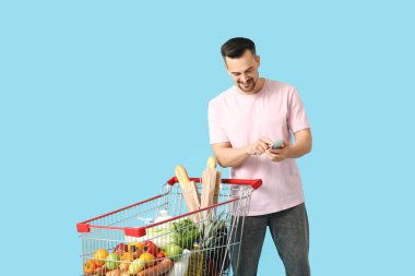 Mavi arka planda tam alışveriş arabası ve cep telefonu olan mutlu genç bir adam.