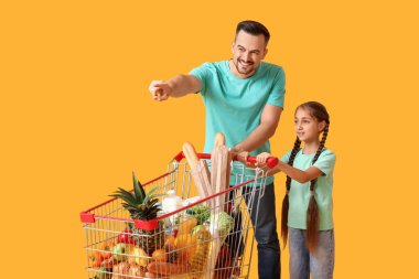 Genç, mutlu baba ve kızı alışveriş arabasıyla sarı arka plandaki bir şeyi işaret ediyorlar.