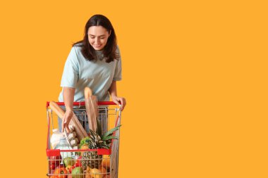 Sarı arka planda alışveriş arabası olan güzel, genç ve mutlu bir kadın.