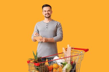 Sarı arka planda alışveriş arabası ve defteri olan mutlu genç bir adam.