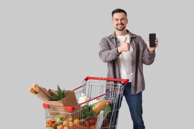 Alışveriş arabalı ve cep telefonlu genç mutlu adam gri arka planda baş parmak hareketi gösteriyor.