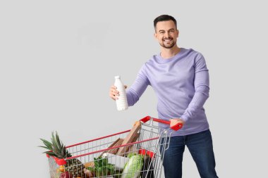 Alışveriş arabası ve gri arka planda bir şişe sütü olan genç ve mutlu bir adam.