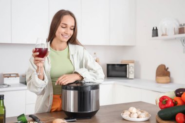Mutfaktaki masada elinde bir bardak şarap ve çoklu tencereyle genç bir kadın.