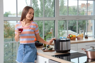 Mutfaktaki masada elinde bir bardak şarap ve çoklu tencereyle genç bir kadın.