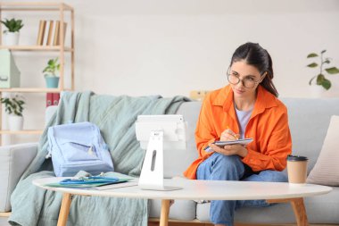 Tablet bilgisayarı, not defteri ve bir fincan kahveyle evde online konferans dinleyen genç bir tıp öğrencisi.