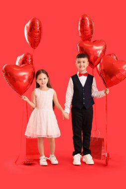 Kırmızı arka planda el ele tutuşan kalp şeklinde balonları olan şirin çocuklar. Sevgililer Günü kutlaması