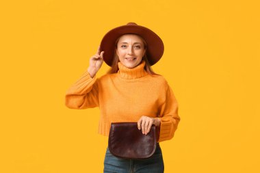 Turuncu süveter ve sarı arka planda kahverengi çantalı şık şapkalı güzel olgun bir kadın.