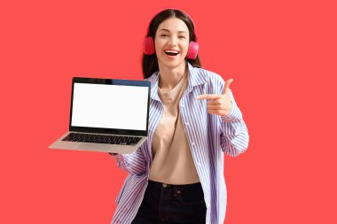 Kulaklıklı güzel bir kadın kırmızı arka planda boş dizüstü bilgisayara bakıyor.