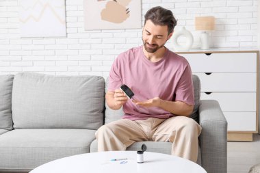 Şeker hastası genç adam evdeki kanepede glukometre kullanıyor.