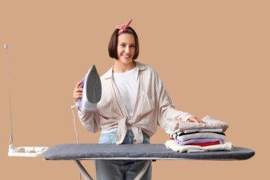 Bej arka planda kıyafet ütüleyen genç bir kadın.