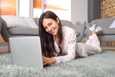 Pembe pijamalı kadın evde dizüstü bilgisayarla halıda çalışıyor.