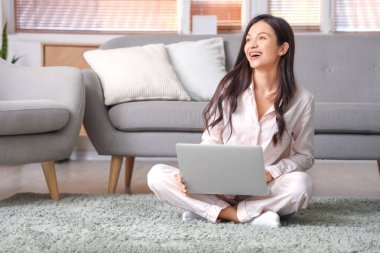 Pembe pijamalı kadın evde dizüstü bilgisayarla halıda çalışıyor.