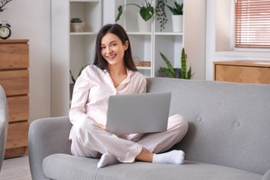 Pembe pijamalı kadın evdeki kanepede dizüstü bilgisayarla çalışıyor.