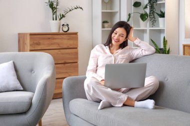 Pembe pijamalı kadın evdeki kanepede dizüstü bilgisayarla çalışıyor.